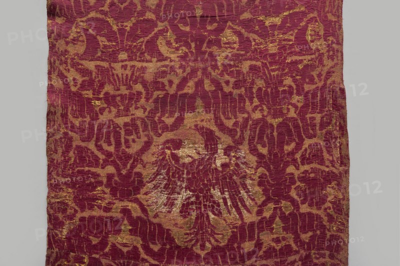 Fragment of fabric, 16th century, Italy. Brocade fragment sewn from several smaller pieces, a ply of fabric with a net composition. In reserves formed by interwoven ribbons are coats of arms Jagiellonian Eagle and bouquets of flowers in vases. Preserved features: patterned fabric with report, preserved ply width and selvedge. Predominant colors: carmine red, yellow, and white; background originally ochre . Height 234 cm, width 56 cm., A rectangular piece of fabric with a repeating ornamental pattern in deep red and muted gold tones, showing a central motif resembling a stylized winged shape formed by the red against the gold background; the textile has an overall textured, worn surface with irregular edges and areas where the gold appears more pronounced or faded, and the pattern consists of flowing, curvilinear shapes and abstract forms distributed across the fabric.