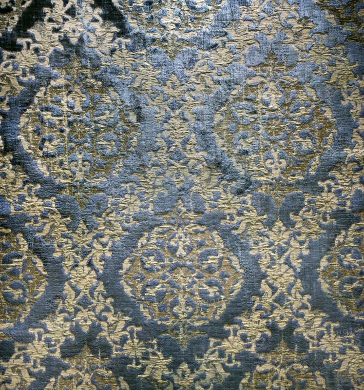 A fabric fragment: a diagonal net of plant and floral tendrils against a blue velvet background. In the fields of the net, in the shape of pointed ovals, are rosettes composed of stylized plant forms. Colors: blue background; ornament sand-colored with gold thread. Materials: velvet and silk; fabrics with preserved selvedge and preserved ply width. Dimensions: fragment 30 x 44 cm; ply width 63 cm; selvedge width 1.3 cm. Date: 15th century, Spain., A close-up of a textured fabric or rug showing an ornate repeating pattern with rounded medallion shapes and intricate floral and scroll-like motifs; the surface has a raised, plush pile contrasting with flatter woven areas, creating visible shadows and highlights. The dominant colors visible are blue and gold with areas where the blue appears darker and the gold ranges from pale to deeper tones. The pattern repeats across the image with symmetrical arrangements and varying pile heights that give a slightly distressed or worn appearance in pl...