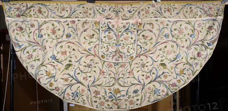 Church cope, Cope made of embroidered silk fabric with linen backing and lining; the original silk backing has crumbled and the embroidery is preserved thanks to an underlying linen backing. Embroidered decoration completely fills the surface with a pattern of leafy tendrils and flowers roses, tulips, carnations. Silhouettes of birds—peacocks, parrots, turkeys, and others—adorn the branches. Edges trimmed with yellow silk braid; hood with preserved bows of pink silk ribbon. 18th century, Silesia. Liturgical vestment. Length 139 cm, width 287 cm., A large semicircular textile with a light cream background covered in an all-over floral and foliate pattern in pastel and muted colors; there are a few shades of pink, blue, green, yellow, and touches of purple and brown in the embroidered flowers, leaves, and curling vines. The fabric shows symmetrical swirling stems and clusters of blossoms and buds, with some small bird shapes worked into the design in green and yellow tones. At the top ed...