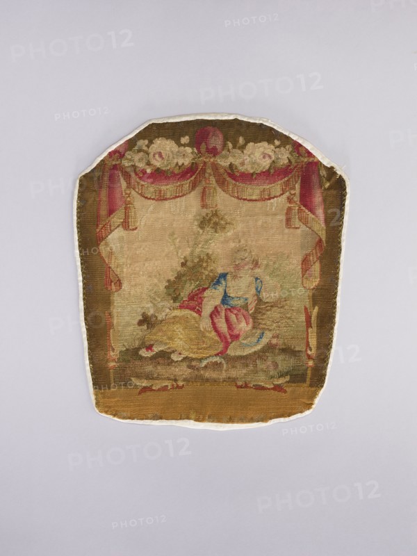 Three tapestry armchair covers, varietals, provenance, flowers, floral ornaments, genre scenes, interior furnishings, garlands, furniture upholstery, decorative fabrics; three tapestries: a boy sitting on a hillock playing a flute, b boy watering flowers, c girl sitting on a hillock; 2nd half of the 18th century; Aubusson; fabrics; height 27.5 cm, width 35 cm., A fabric panel with a rounded top and straight bottom showing a woven scene in muted colors; the panel features a decorative border with draped curtains and floral swags in shades of red, pink, cream, and brown, framing a central pastoral scene. Within the scene a reclining figure wearing garments in blue, pink, and yellow is set against an earthy background of greens, browns, and beige with hints of darker green foliage behind. The overall color palette includes brown, beige, cream, pink, red, blue, yellow, and green, and the panel edges show a lighter fabric backing in off-white.