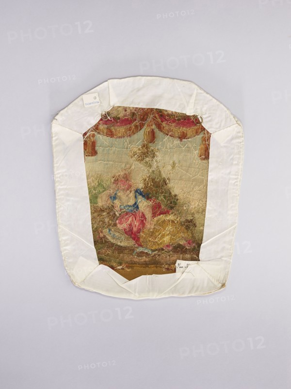 Three tapestry armchair covers, varietals, provenance, motifs: flowers, floral ornaments, genre scenes, interior furnishings, garlands, furniture upholstery, decorative fabrics, tapestries; scenes: a boy sitting on a hillock playing a flute, b boy watering flowers, c girl sitting on a hillock; 2nd half of the 18th century, Aubusson; fabrics; height 27.5 cm, width 35 cm., An irregularly shaped textile piece mounted on a pale background, bordered by a white fabric frame with folded corners; the central embroidered area shows a worn multicolored scene with a dominant palette of muted gold, beige, red, blue, green, and brown, including a group of soft, frayed thread forms suggesting figures and drapery, a decorative scalloped valance in red and gold tones along the upper edge, and areas of loose, tangled fibers and faded stitching across the surface.