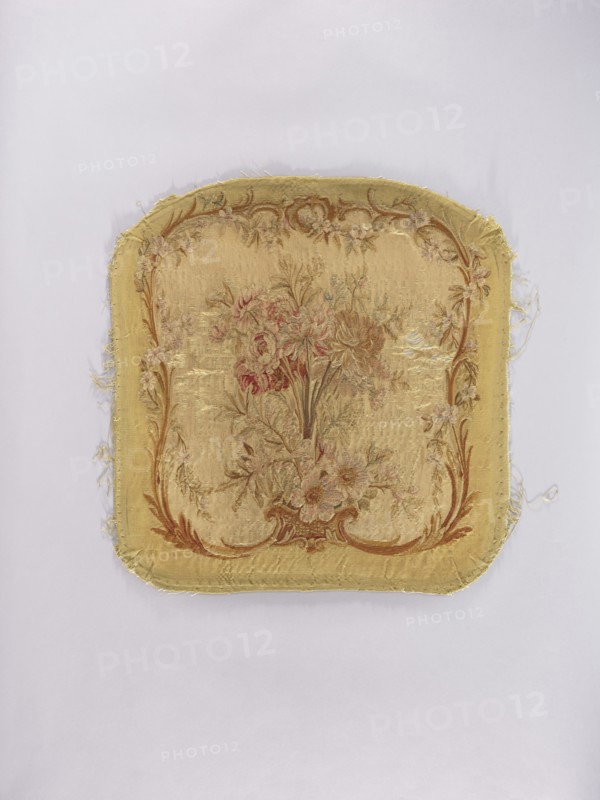 Two tapestry armchair backrest upholsteries and one sofa upholstery, unknown author, varietal, flowers, floral ornaments, furniture upholstery, interior furnishings, decorative fabrics, tapestries; a, b armchair upholstery with a bouquet of flowers in the center, surrounded by a frame of acanthus flowers and leaves; c sofa upholstery with a spreading bouquet of flowers surrounded by a frame with acanthus flower and leaf motifs. Colors: sandy background, olive, pink, red, ochre, blue, and green ornaments. 18th/19th century, France; fabrics; height 64 cm, width 62 cm., A square textile fragment with rounded corners lying on a pale background, showing a woven floral central design in faded tones of gold, beige, pink, green, and brown, surrounded by a decorative border in similar gold and brown shades; the fabric edges are frayed and worn, with visible stitched seams and areas where threads have come loose, and the surface displays subtle variations in sheen and some worn patches.
