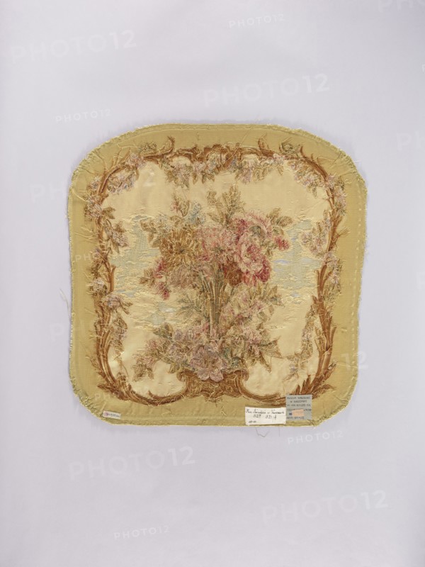 Two tapestry armchair backrest upholsteries and one sofa upholstery, varietal, flowers, floral ornaments, furniture upholstery, interior furnishings, decorative fabrics, tapestries; a, b armchair upholstery with a bouquet of flowers in the center, surrounded by a frame of acanthus flowers and leaves; c sofa upholstery with a spreading bouquet of flowers surrounded by a frame with acanthus flower and leaf motifs. Colors: sandy background, olive, pink, red, ochre, blue, and green ornaments. 18th/19th century, France; fabrics; height 64 cm, width 62 cm., A square textile with rounded corners is centered on a pale background; the textile is primarily yellow and beige with frayed edges and a decorative woven floral scene in the center showing a bouquet of flowers in shades of pink, red, brown, green, and muted blue, surrounded by an ornate border in brown and gold tones with additional small floral motifs, and a few small rectangular tags attached near the lower edge.