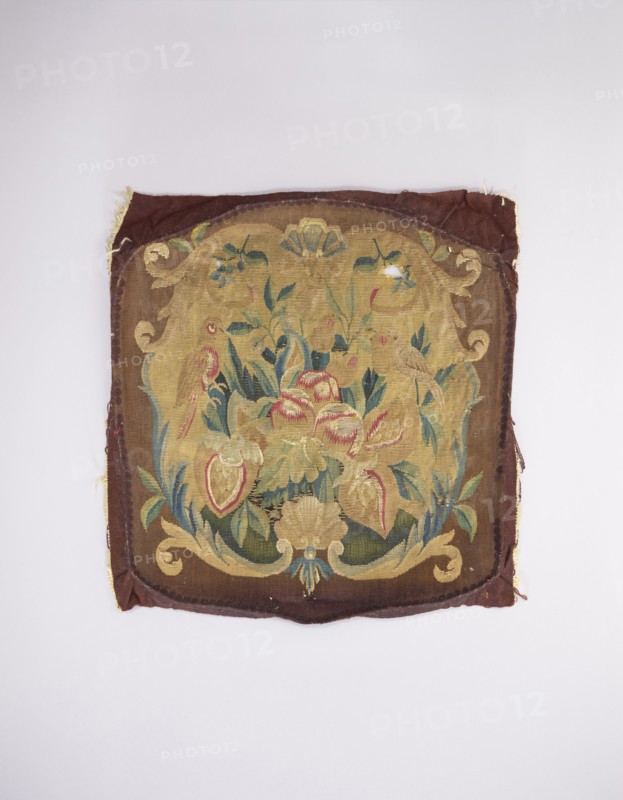 Tapestry upholstery for the seat and backrest of an armchair, unknown author, unknown provenance; flowers, floral ornaments, shells, birds; interior furnishings, furniture upholstery, decorative fabric tapestry; a seat, b backrest. Surrounded by a frame with motifs of acanthus leaves, shells, and flowers. Colors: brown and yellow background, red, dirty pink, sky blue, green, and yellow-green ornament. Late 18th century, France. Fabrics; height 59 cm, width 57 cm., A square textile with a worn, decorative woven panel featuring a central floral arrangement framed by a scrolling cartouche; the fabric shows frayed edges and a few small holes. The visible colors include shades of brown and tan in the background and border, muted gold and yellow tones in the central cartouche, and green and blue hues in the leaves and foliage. There are accents of red and pink in some of the flower forms and pale beige in other floral elements, along with small areas of darker brown and olive. The piece rest...