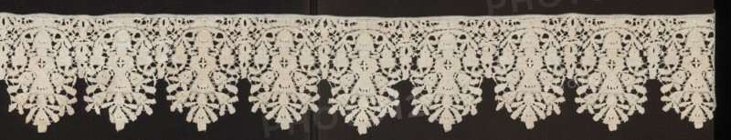 Lace fragment, floral ornaments. Lower edge serrated. In each oval there is a vase of flowers. Flower crowns overlap the serrations. Joined with spiderwebs, so-called Van Dyck. Date: 1630–1650. Place of origin: Flanders. Material: lace. Dimensions: length 200 cm, width 13.5 cm., A horizontal strip of lace with a repeating scalloped pattern is visible against a dark background; the lace is a pale off-white or cream color and features a series of arch-like motifs connected along a straight top edge, each arch enclosing a symmetrical floral or fern-like pendant that hangs down with layered, leaf-shaped elements and openwork details, and the dark background shows through the lace openings creating contrast between the light lace and the deep dark backdrop.