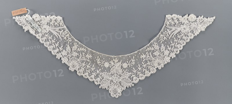 Collar, bobbin lace, 19th century, Europe; women's clothing accessory; width 11 cm, length 34.5 cm. Floral ornament includes various flower rosettes: small 5-leaf rosettes, larger feathery ones at the corners, bell-shaped calyxes with a stripe on the edges, small lanceolate and larger divided leaves., A delicate triangular lace collar displayed flat against a gray background, made of off-white lace with a fine mesh ground and an ornate floral pattern; the scalloped edge is lined with embroidered flowers and leaves, with several larger floral motifs toward the center and smaller sprigs and dotted mesh filling the interior, creating an overall symmetrical, lacy appearance with intricate needlework and a subtle contrast between the off-white lace and the smooth gray backdrop.