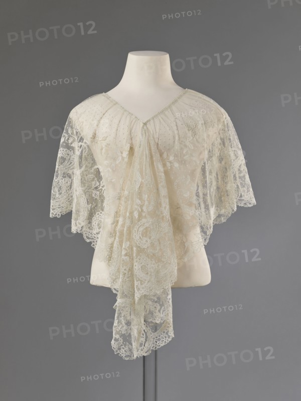 Collar, lace collar so-called berta with a pattern of flowers, leaves and small dots in the background; floral and plant ornaments; clothing accessory for women's fashion; 1840–1860; Brussels, lace; width 33 cm., A mannequin torso displays a delicate lace shawl or capelet in shades of cream and off-white draped over its shoulders, with sheer floral lace panels and scalloped edges that hang down in a layered, flowing arrangement; the mannequin is white and mounted on a thin stand against a smooth gray background.