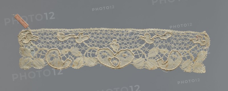 Cuff, cuffs; fashion accessory; lace. Pattern of leaves and palmettes at the lower edge. Mesh background with triangular and round eyelets. Straight upper edge. 2nd half of the 19th century, Germany . Clothing accessory; width 6 cm, length 29 cm., A narrow strip of delicate lace in shades of off-white and cream is set against a solid gray background; the lace has an open, net-like upper section with a decorative lower border featuring a repeating pattern of heart-like swirls and leaf shapes, and a few raised, corded outlines that add texture; small irregularities and slight discolorations in pale brown and tan appear in places along the lace, and a small pinkish ribbon is attached near one end.
