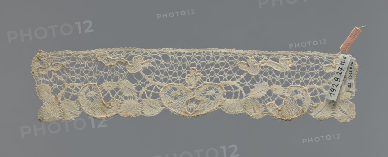 Cuff, repossession provenance; fashion accessory cuff; lace; pattern of leaves and palmettes at the lower edge; mesh background with triangular and round eyelets; straight upper edge; 2nd half of the 19th century; Germany . Dimensions: width 6 cm, length 29 cm., A narrow strip of lace with an openwork pattern laid flat against a neutral gray background, showing a scalloped lower edge with curved, leaflike and teardrop motifs and an upper edge with a more irregular honeycomb-like mesh; the lace appears off-white to cream with areas of slightly darker beige tones and small accents of pale brown discoloration, and there is a small fabric tag attached at one end in a muted pinkish color with a small pale cloth label nearby.