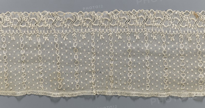 Lace fragment, Alençon lace, lace; plant ornaments. At the bottom, an edging ornament running in a clearly separated strip, with plant motifs and an openwork form resembling the letter S repeated regularly. Higher on the mesh, oval stripes between which projections: small diameter points. 1st half of the 18th century, France. Material: fabric. Width 17 cm, length 206 cm., A horizontal strip of delicate lace fabric in a pale cream color lies against a muted gray background, showing an upper scalloped edge with ornate looping floral motifs and a lower straight edge with a finer scalloped trim; the main body of the lace displays a repeating vertical pattern of open oval chain-like columns separated by mesh areas dotted with small raised bobbles, and the overall texture appears lightweight and finely netted with subtle variations in thread thickness and tiny decorative loops along the borders.
