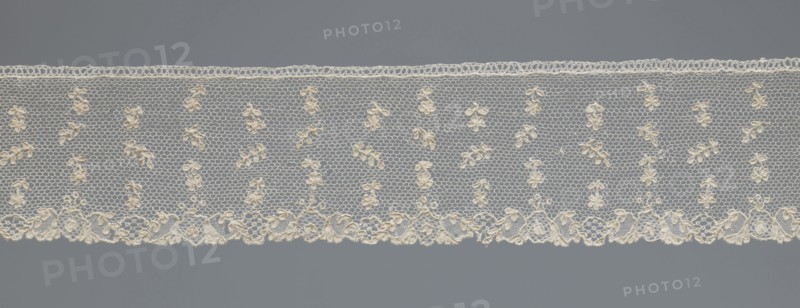 Fragment of lace;, One-sided strip of edge ornament made of plant forms: motif of a pinnately leafed twig. Above the edge ornament, against a mesh background in projections, an alternating twig with a single flower and a pinnately leafed twig. 18th century, France. Lace; width 6 cm, length 63 cm., A horizontal strip of delicate lace displayed against a uniform background, showing a repeating hexagonal net pattern with small floral motifs embroidered throughout and a more elaborate scalloped border along one edge; the lace appears in shades of off-white and cream while the background is a muted gray.