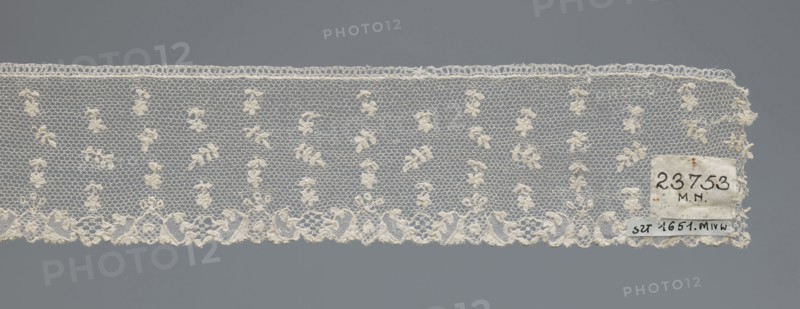 Fragment of lace, one-sided strip of edge ornament made of plant forms with a motif of a pinnately leafed twig; above the edge ornament, against a mesh background in projections, an alternating twig with a single flower and a pinnately leafed twig. Lace, 18th century, France. Width 6 cm, length 63 cm., ., A horizontal length of delicate lace displayed against a neutral gray background, showing a fine hexagonal net ground with a repeated motif of small clustered floral-like knots in an off-white or cream color, a scalloped lower edge with denser embroidered floral and scroll designs, a straight upper edge with a narrow looped border, and a small rectangular patch of fabric attached near one end in similar cream tones.