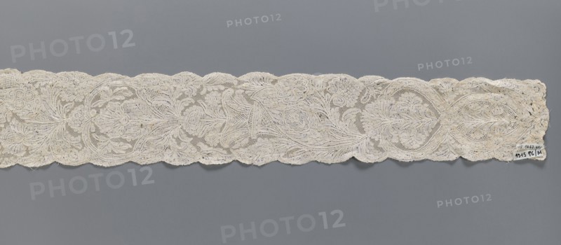 Lace fragment, Binche lace, plant ornaments, floral ornaments. Floral and plant pattern in a candelabra arrangement, with flowers enclosed in an oval at the bottom and freely drawn at the top. Mesh background of the ißsgrond and point de neige types. Ca. 1750. Lace, width 10 cm, length 118 cm., A long narrow piece of lace with a scalloped top and bottom edge stretches across the image against a solid gray background, showing an intricate floral pattern with flowing vine-like motifs and open mesh areas; the lace appears in shades of off-white, cream, and light beige with subtle variations in tone, and there is a small irregularity at one end where the fabric looks slightly frayed.