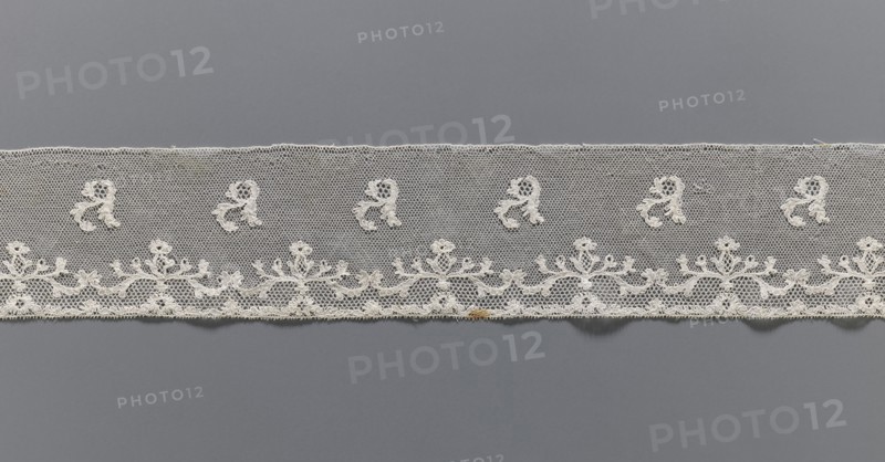Lace fragment, Plant ornaments, Louis XVI style. At the lower edge there are two trails of fine tendrils, and between them a net with issgrond, mesh. Above, a net with drochelle mesh, on it projections of leafy branches. Contours of motifs reinforced with thread. Late 18th century, Mechelen. Malines lace. Lace, width 7.5 cm, length 146 cm., A horizontal strip of delicate lace in off-white stretched across a smooth gray background, featuring a netted upper field with a row of small ornamental motifs and a lower scalloped border with an ornate floral-like embroidered pattern, the lace showing fine openwork, looping threads, and a small area with a darker ochre discoloration near the bottom edge.