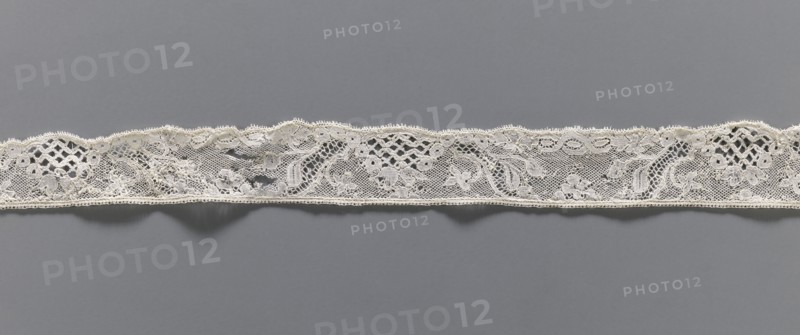 Lace fragment, floral ornaments, Malines lace, pattern with motifs of winding ribbons, flowers, and rococo elements; Point de drochelle background. 2nd half of the 18th century, Mechelen. Lace, width 3.5 cm, length 83 cm., A narrow strip of delicate lace runs horizontally across a flat gray background, showing an intricate openwork pattern with floral and leaf motifs, scalloped upper edge and a straighter lower edge, featuring areas of fine netting, looped decorative borders, and denser embroidered sections; the lace is primarily off-white or cream against a medium gray backdrop.