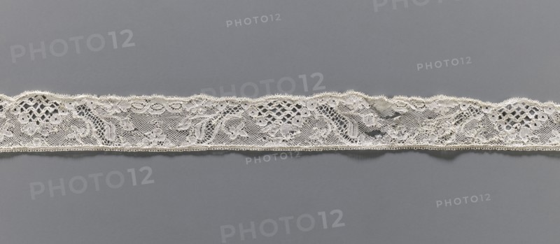 Lace fragment, Floral ornaments. Malines lace. Pattern with motifs of winding ribbons, flowers, and rococo elements. Point de drochelle background. 2nd half of the 18th century. Mechelen. Lace, width 3.5 cm, length 83 cm., A horizontal strip of delicate lace with floral and geometric openwork motifs lies across a smooth background; the lace is an off-white or cream color with intricate netted areas and scalloped top edge, and the background is a uniform medium gray.