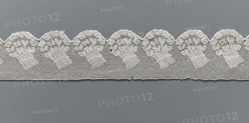 Fragment of lace, Malines Mechelen lace, 18th century, from Henryków Heinrichau, lace, width 6 cm, length 62 cm, A strip of delicate lace with a scalloped top edge lies horizontally against a uniform gray background; the lace is off-white and features a repeating pattern of ornate floral bouquets worked into a fine hexagonal mesh, with the scallops forming rounded arches above each bouquet and a straight lower edge below; the floral motifs show clustered rounded shapes and textured stems, and the lace displays subtle variations in thread density and tiny openings where the mesh is visible.