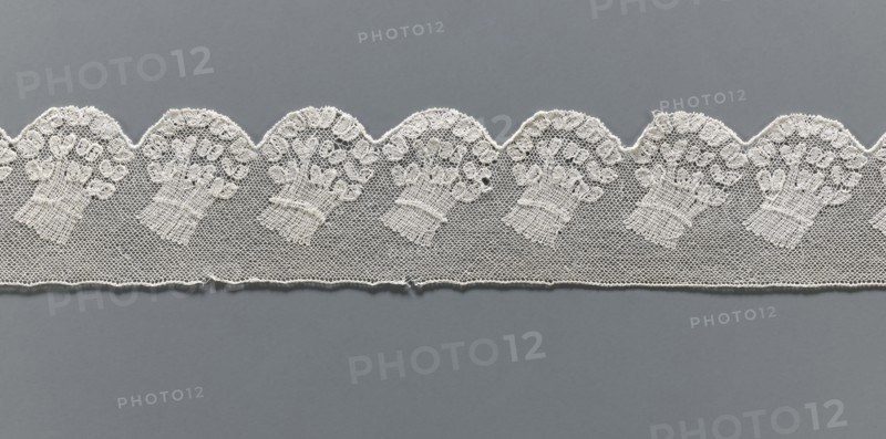 Fragment of lace, Malines Mechelen lace, 18th century, Henryków German: Heinrichau, lace, width 6 cm, length 62 cm., A horizontal strip of delicate white lace with a scalloped upper edge rests against a muted gray background; the lace features a repeating floral bouquet motif worked in fine thread over a hexagonal net ground, showing a row of scallops with ornate flower clusters and textured stems, and the lower edge of the lace has a subtle finished border with a few small loose threads. The visible colors are white in the lace and gray in the background.