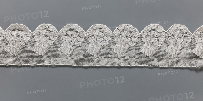 Fragment of lace, Malines Mechelen lace, 18th century, Henryków Heinrichau, lace, width 6 cm, length 33 cm, A strip of delicate white lace with a scalloped upper edge lies horizontally across a flat gray background, the lace featuring a repeating motif of bouquet-like floral shapes worked into a fine netted mesh; the visible colors are white and gray.