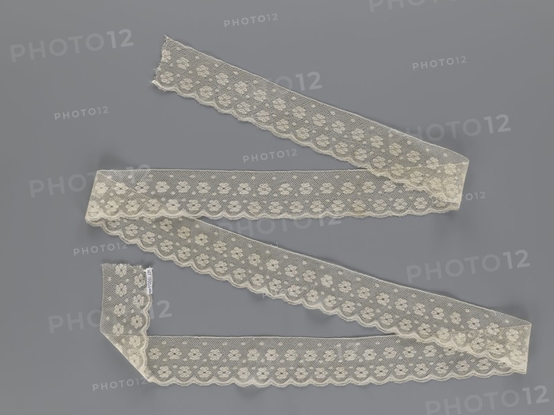Lace fragment, tied in the form of a belt with one-sided arched serrations. Regular ornament: rosettes and points arranged in rows. Mid-19th century, possibly Flanders. Lace, width 7 cm, length 223.5 cm. Maker: Kublicki Piottuch, Alfred 1861–1945., A length of scalloped lace ribbon arranged in a zigzag pattern on a flat surface, showing a delicate floral mesh design with a scalloped edge; the lace is an off-white or cream color and the background is gray.