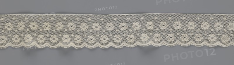 Lace fragment, tied in the form of a belt with one-sided arched serrations. Regular ornament: rosettes and points arranged in rows. Mid-19th century, Flanders, lace, width 7 cm, length 223.5 cm., A horizontal strip of delicate lace is shown against a flat gray background, with the lace in an off-white or cream color featuring a repeating pattern of floral motifs connected by a net-like mesh and small circular accents, the lower edge forming a scalloped border with tiny looped fringes, and the overall texture appearing fine and intricate.