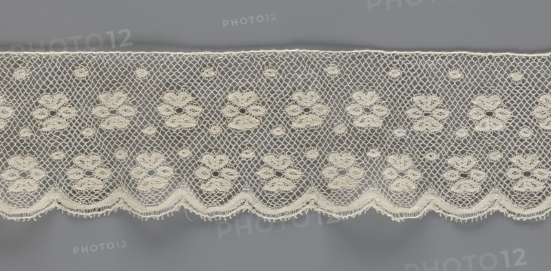 Lace fragment, tied in the form of a belt with one-sided arched serrations. Regular ornament: rosettes and points arranged in rows. Mid-19th century, possibly Flanders. Lace, width 7 cm, length 223.5 cm., A horizontal strip of delicate lace in off-white with a repeating floral motif set against a gray background, featuring a net-like mesh between the floral clusters, small circular lace accents scattered throughout, and a scalloped lower edge with textured fringe; colors visible include off-white and gray.