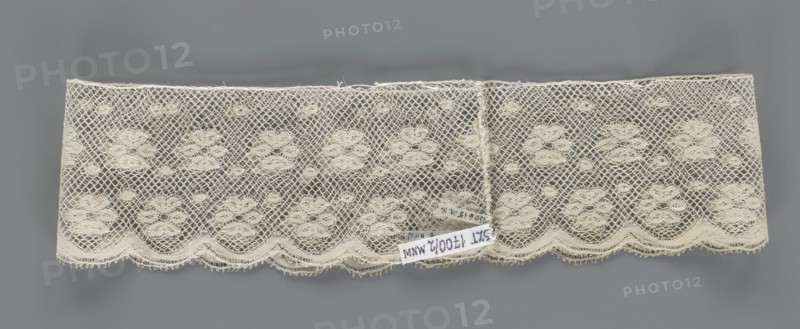 Fragment of lace, a section of a belt sewn into a closed circle with one-sided arched serrations. Regular ornament: rosettes and points arranged in rows. Mid-19th century, Flanders, lace, width 7 cm, length 50 cm., A rectangular piece of delicate lace with an open netted pattern and floral motifs, laid flat against a gray background; the lace is a creamy off-white color with a scalloped lower edge and a few small loose threads along the top; the netting forms diamond-shaped holes surrounding clusters of rounded floral shapes, and there is a small group of labels attached near the lower center that contrast with the lace.