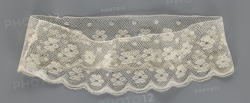 Fragment of lace, lace. A section of a belt sewn into a closed circle with one-sided arched serrations. Regular ornament: rosettes and points arranged in rows. Mid-19th century, possibly Flanders. Lace, width 7 cm, length 50 cm., A length of delicate lace fabric displayed against a neutral background, showing an open mesh pattern with a repeating floral motif and scalloped edge; the lace appears in off-white or cream tones while the background is a medium gray, and the fabric has slight folds and shadowing that create areas of lighter and darker cream.