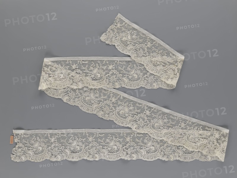 Lace fragment, lace, floral ornaments with rocaille motifs, over-stylized fan-shaped ribbons with arcades, twigs, dotted background, point de gaze ground. Lower edge serrated to match motifs; upper edge straight with a bobbin insert. Some rose flowers are in relief with petals made separately and attached by one edge. 1st half of the 19th century, France, lace, width 15 cm, length 298 cm, A length of delicate lace ribbon arranged in gentle S-shaped curves against a smooth background, the lace displaying an intricate floral and leafy pattern with scalloped edges and fine netting, the visible colors being off-white for the lace and a muted gray for the background.