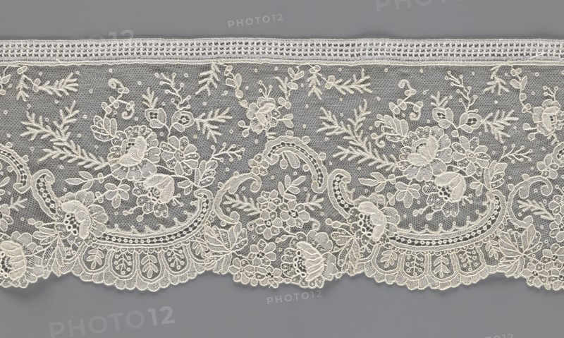 Lace fragment, lace, floral ornaments with rocaille motifs, over-stylized fan-shaped ribbons with arcades, twigs, dotted background, point de gaze background. Lower edge serrated to match motifs; upper edge straight with a bobbin insert. Some rose flowers formed in relief with petals separately made and attached by one edge. 1st half of the 19th century, France, width 15 cm, length 298 cm., The image shows a length of delicate lace with a floral and scroll pattern against a gray background; the lace is ivory or off-white and features a fine net ground dotted with small picot-like points, a scalloped lower edge with ornate motifs, and an upper straight edge with a narrow band; the design includes a group of stylized flowers, leaves, buds, and curling vines arranged in repeating panels, with varying stitch textures creating raised and open areas; visible colors are gray for the background and ivory or off-white for the lace.