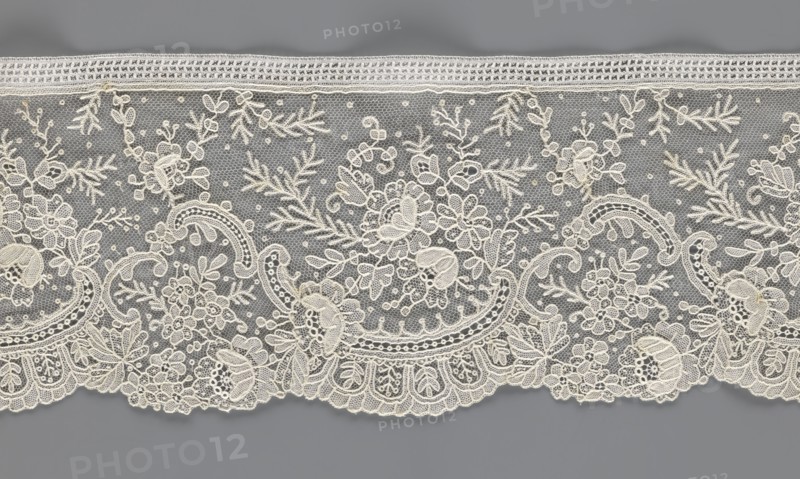 Lace fragment, lace with floral ornaments, rocaille motifs, over-stylized fan-shaped ribbons with arcades, and twigs; dots in the background; point de gaze background; lower edge serrated to match motifs, upper edge straight with a bobbin insert; some rose flowers worked in relief with petals made separately and attached by one edge; 1st half of the 19th century; France probable; lace; width 15 cm, length 298 cm., A length of delicate lace with an intricate floral and foliate pattern laid against a neutral background, showing a scalloped lower edge and a straight upper border; the lace features a fine hexagonal mesh filled with a group of embroidered flowers, leaves, scrolls, and small dot motifs, with emphasized outlines and textured fill stitches creating raised details; visible colors include off-white or cream embroidery atop a medium gray background, with subtle variations in the cream threads giving a slightly warm tone against the cool gray.