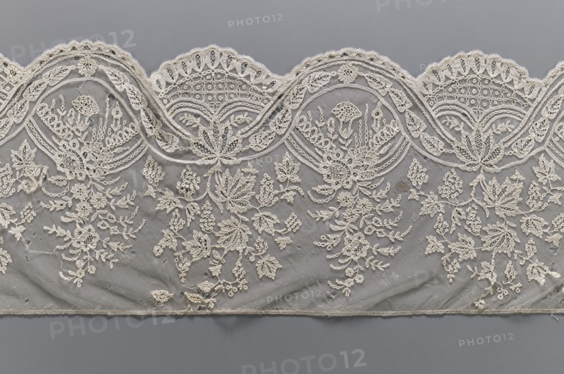Lace fragment, bobbin lace, mid-19th century, Brussels. Two ribbons: one wider with leaves, the other narrower with a chain; interlacing to create ovals containing garlands. Branches with flowers or vine branches with fruit extend upward from the ovals. Background in fond de gaze. Lower edge semicircularly serrated, upper edge straight. Materials: lace. Width 28 cm, length 187 cm., A scalloped piece of lacework with an intricate floral and leaf embroidered pattern laid against a smooth background; the lace is a creamy off-white with detailed loops, mesh netting, and raised stitched motifs forming bouquets, sprigs, and clustered blossoms, while the background is a muted gray that contrasts with the lace; the lace edge shows repeating rounded arches with decorative openwork and stitched borders, and the lower edge finishes with a narrow stitched hem.