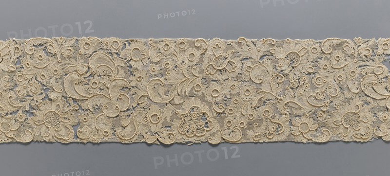 Fragment of lace, lace, plant ornaments, ca. 1700, France, lace, width 17 cm, length 157 cm, A long strip of intricately patterned beige lace laid horizontally against a smooth gray background, featuring a dense floral and foliate design with a few large stylized blossoms, several smaller rosette-like motifs, and many curving paisley-shaped elements; the lace shows varied openwork areas and raised embroidered outlines, with visible texture from delicate netting, looping threads, and tiny picot edges, and the visible colors are beige and gray.