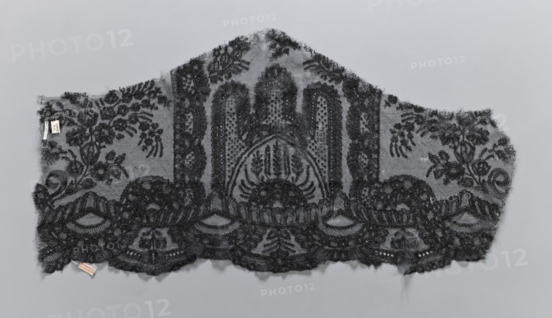 Lace fragment, plant ornaments. Preserved ruffle fragment features a symmetrical ornament of a central arch with three pinnacles. Serrated edge decorated with a wide band of edge ornament composed of arch segments with geometric ornament and an interwoven tendril from a band of plant-geometric ornament. 19th century, France. Lace, width 38 cm, length 74 cm., A scalloped piece of delicate black lace fabric with an ornate floral and foliate pattern laid flat against a neutral gray background; the lace shows a central arched motif with dense embroidered detailing and dotted netting, flanked by symmetrical botanical sprays and scrolling leaves, and a lower scalloped border of dense floral embroidery and openwork; loose threads and small frayed edges are visible along the outer edges, and a small fabric tag is attached near one corner.