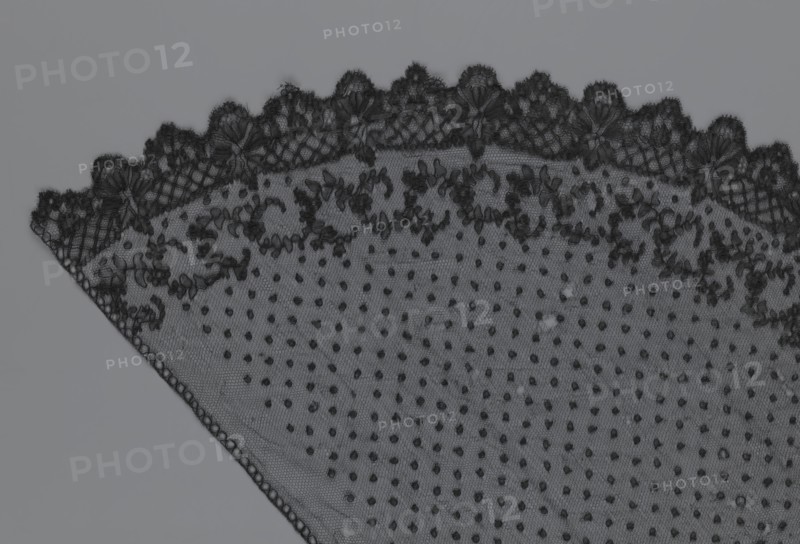 Veil, veils, lace, clothing accessories, fashion. Lace with a floral-geometric pattern running in a wide strip along the semicircular, serrated edge. The remaining part features fond de Tull and regularly spaced small circles. 19th century, France. Clothing; height 35 cm, width 74 cm., A triangular piece of lace fabric occupies the image, showing a scalloped upper edge with intricate embroidered floral motifs and a fine netting background; the main field of the fabric features a dotted pattern of small raised tufts, and there is a narrow woven border along one slanted edge. The visible colors are shades of black and gray.