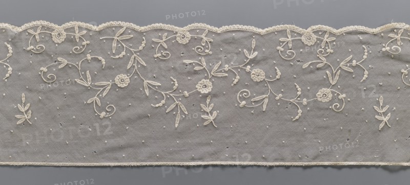 Lace fragment, lace, plant ornaments. The lower edge is gently serrated with flat arches, bordered with a band of fine, bobbin-shaped teeth, looped onto the mesh. Ornament elements made with chain and bobbin-shaped techniques. Bobbin-shaped mesh, hand-made. 2nd half of the 19th century, Europe. Lace, width 26.5 cm, length 283 cm., A strip of delicate net fabric with an ornate scalloped edge is shown against a gray background; the net is a light cream color and features embroidered floral and leafy motifs in the same cream tone that form flowing, curling patterns, with a few small raised dots scattered across the surface; the scalloped border has a lace trim and the lower edge shows a narrow plain hem, while the background behind the net appears as a uniform medium gray.