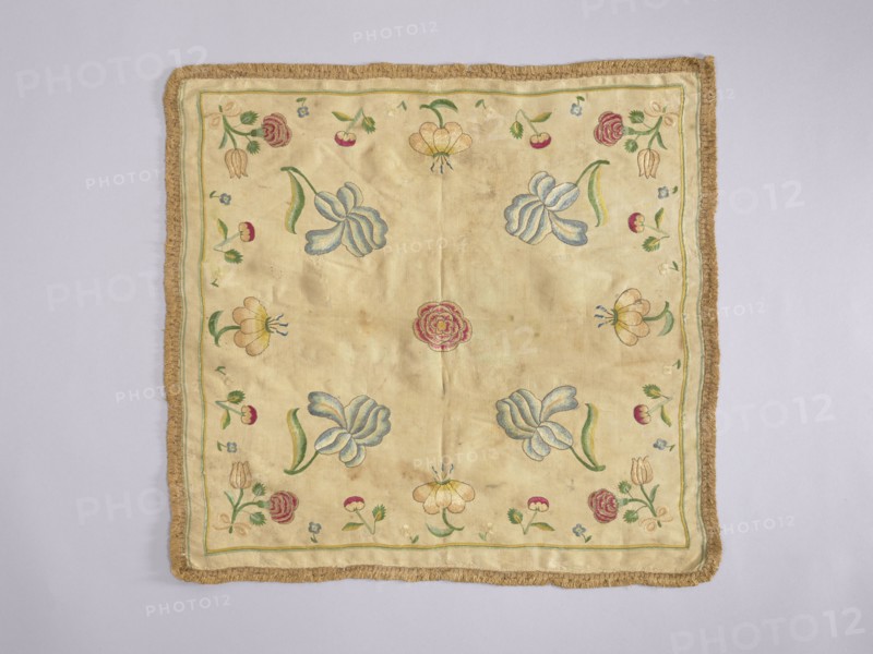 Church napkin, Church paraments; flowers; leaves; plant ornaments; sacred art. Napkin of white silk fabric embroidered with single projections of large flowers and fruit. Trimmed with silk braid with short shreds. Plain weave fabric, density 34 weft × 34 warp threads per cm, two workshop edges preserved, piece width 45 cm. Flat, pierced embroidery with colored silk threads — a thread of one color penetrates a row of threads of another color creating shaded transitions. Embroidery with gold thread around petals of peonies, irises, and tulips. Colors subdued but not faded: burgundy, multiple blues, cream, beige, ochre, green, light yellow-green, gold. Compared to a related napkin, colors are less bright and embroidery more detailed; napkin also appears earlier. Early 18th century, Poland. Height 42.5 cm, width 45 cm., A square textile with a fringe border in a golden brown color lies flat against a light gray background; the central field of the textile is a pale beige or cream tone embr...