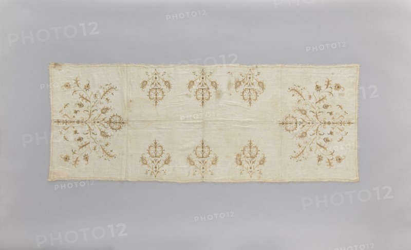 Church napkin of thin white linen 25 warp and weft threads per cm covered with embroidery featuring a pattern of large, evenly distributed, stylized Eastern flower bouquets. Flat, double-sided, pierced embroidery, using silk and gold thread. Very fine stitches. Cream and gold predominate in the ornament. Edged with cream veining. All edges with warp or weft threads ripped out to obtain a short fringe, approx. 1 cm long. 17th/18th century, Poland. Height 174 cm, width 65.5 cm. Subject: flowers, leaves, plant ornaments; sacred art; used as church paraments in Evangelical churches. Materials: linen, silk, gold thread., A rectangular fabric panel with a light beige background displayed flat against a pale gray backdrop, featuring symmetrical ornamental motifs in shades of brown and tan arranged across the surface; the cloth shows subtle wrinkles and a slightly frayed edge, with a few darker brown spots and stains visible, and several ornate floral and scroll-like designs positioned along t...