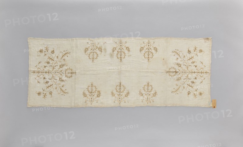 Church napkin of thin white linen 25 warp and weft threads per cm covered with embroidery featuring a pattern of large, evenly distributed, stylized Eastern flower bouquets. Flat, double-sided, pierced embroidery, using silk and gold thread. Very fine stitches. Cream and gold predominate in the ornament. Edged with cream veining. All edges with warp or weft threads ripped out to obtain a short fringe, approx. 1 cm long. 17th/18th century, Poland. Materials: linen, silk, gold thread. Height 174 cm, width 65.5 cm. Function: church napkin; motif: flowers, leaves, plant ornaments; sacred art; used in churches and chapels., A rectangular textile laid flat on a neutral background showing a pale cream or off-white base with decorative floral motifs in a muted tan or light brown. The fabric has frayed edges and visible creases and folds. Symmetrical ornate floral arrangements appear along the length, with a few larger, branching spray designs near the shorter ends and several smaller, repeatin...