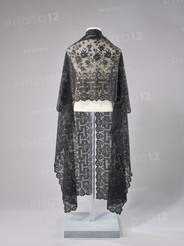 Shawl, shawl; 1st half of the 19th century, France; women's clothing; clothing accessory; floral ornaments with meander motif — the meander is associated with straight flower branches with a single large flower directed towards the outer edge; outer edge serrated; lace; length 270 cm; width 63.5 cm; provenance: purchase., A mannequin draped in a long, sheer lace garment with floral and geometric patterns, shown from the back against a neutral backdrop; the lace is dark and contrasts with a light underlayer around the shoulders and waist, and the garment falls into scalloped edges with decorative motifs; the base and support are visible beneath the fabric and the background is a smooth gradient of pale gray.