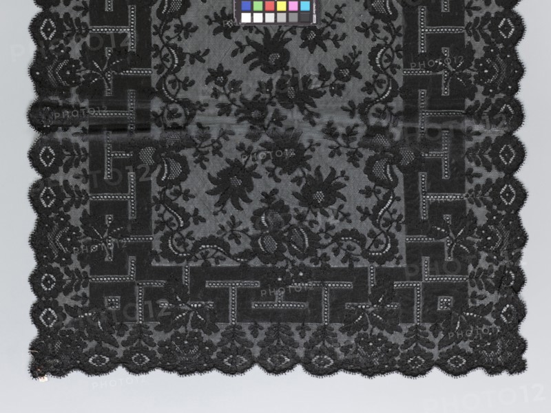 Shawl, shawl; shawls; fashion; women's clothing; clothing accessories; floral ornaments; meander ornament. The meander is associated with straight flower branches with a single large flower directed towards the outer edge. The outer edge is serrated. 1st half of the 19th century, France. Clothing, lace. Length 270 cm, width 63.5 cm., A rectangular piece of dark lace fabric with an ornate floral and geometric pattern occupies most of the image, showing a scalloped edge along the bottom and sides; the lace is primarily black with variations in shading where the intricate floral motifs, leaves, and vine-like scrolls contrast against a slightly lighter background, and a border of blocky geometric shapes alternates with floral elements around the interior; a small color calibration chart with several colored squares appears near the top edge, and the overall background behind the lace is a pale gray.