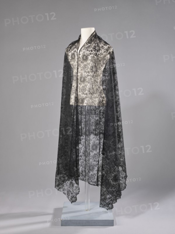 Shawl, shawl, floral ornaments, clothing accessory for women, fashion. Pattern on a mesh background. Long edges: upper edge sewn-on narrow stripe made using the bobbin technique; lower edge serrated without distinctive ornament. Short sides cut off and left unfinished. 1st half of the 19th century, France, fabrics. Dimensions: length 245 cm, width 100 cm., A mannequin draped with a long, sheer black lace shawl over a light-colored sleeveless top with a floral lace pattern; the shawl falls to the floor and has scalloped edges, creating a layered translucent effect that reveals the floral motifs beneath. The mannequin is displayed against a neutral gray background and stands on a pale rectangular platform with a clear vertical support visible beneath the garment. The visible colors are black, light cream or off-white, gray, and pale blue on the platform.