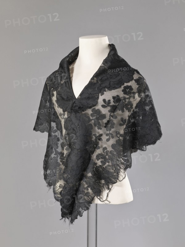 Cape, capes, fashion accessories, fashion, women's clothing. Cape in the form of an elongated rhombus, made of black lace with floral motifs. The edge is serrated in accordance with the forms of floral rosettes placed in a row along the edge. 2nd half of the 19th century, France. Garment, height 75 cm, width 164 cm., A dress form draped with a delicate black lace shawl featuring a floral pattern; the lace is sheer in places revealing the pale cream color of the form beneath, and the edges of the shawl are slightly frayed with a scalloped finish. The background is a smooth gray. The visible colors are black, cream, and gray.