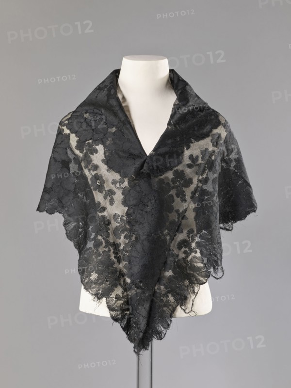 Cape in the form of an elongated rhombus, made of black lace with floral motifs. The edge is serrated in accordance with the forms of floral rosettes placed in a row along the edge. 2nd half of the 19th century, France. Garment; height 75 cm, width 164 cm., A dress form wearing a sheer black lace triangular shawl draped over the shoulders, with floral lace motifs and scalloped edges, the lace appearing delicate and slightly frayed in places, set against a plain light gray background; visible colors include black, off-white from the dress form, and shades of gray in the background.