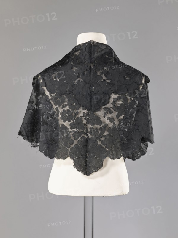 Cape, capes, fashion, women's clothing. Cape in the form of an elongated rhombus, made of black lace with floral motifs. The edge is serrated in accordance with the forms of floral rosettes placed in a row along the edge. 2nd half of the 19th century, France. Garment. Height 75 cm, width 164 cm., A black lace shawl with a floral pattern draped over a white mannequin torso against a neutral gray background, showing scalloped edges and sheer areas where the mannequin is visible; colors visible include black, white, and shades of gray.