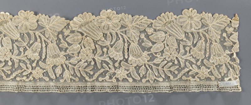 Lace fragment, floral ornaments; fragment of bobbin lace with relief edges made with a needle on a tulle background, featuring a motif of large flowers rosettes and bells and leaves; 19th century, Brussels; lace fabric, width 15 cm, length 110 cm., A wide piece of lace fabric with an overall cream color set against a gray background, featuring a dense floral and leafy pattern with several large scalloped flower motifs along the top edge and a group of bell-shaped blossoms and smaller flowers dispersed throughout; the lace has a fine netted ground with raised embroidered outlines and textured filling stitches creating petals, leaves, stems, and small round accents, and a horizontal decorative border along the bottom edge with open rectangular mesh and a narrow scalloped trim.