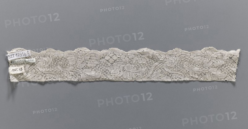 Lace fragment, bobbin lace, white, with a floral tendril ornament on a point de neige background, ca. mid-18th century, Binche possible origin, fabrics, width 4.5 cm, length 32 cm., A long band of lace in off-white is displayed horizontally against a muted gray background, showing an intricate openwork pattern with floral and scroll-like motifs and a scalloped upper edge; the lace appears delicate with fine netting between the motifs and a slightly irregular lower edge, and there is a small cluster of fabric tags attached near one end in off-white and pale beige tones.