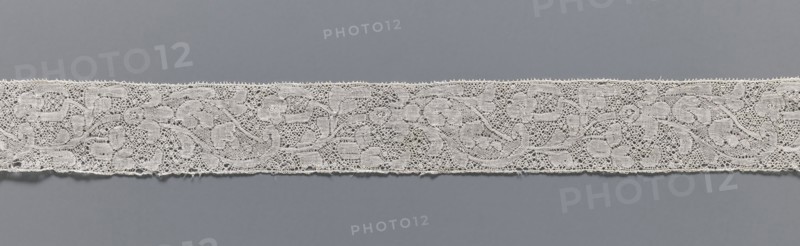 Lace fragment, lace, Binche lace, Henryków German: Heinrichau, clothing accessory, needle lace with point de neige background, straight edges, mid-18th century, Binche, width 3.5 cm, length 37 cm, A horizontal strip of delicate lace in an off-white or cream color runs across a smooth, muted gray background; the lace features a repeating floral and vine pattern with openwork areas and fine netting, scalloped tiny loops along the top and bottom edges, and a few small loose threads visible along the lower edge, while the background shows subtle variations of gray.