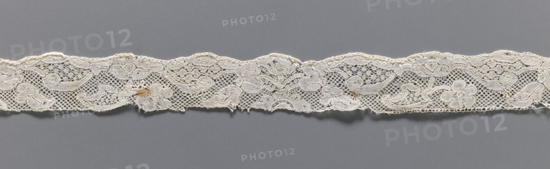 Lace fragment, repossession provenance; bobbin lace, Malines Mechelen style, mid-18th century; fashion accessory. Motifs: floral sprigs, palmettes, leafy tendril, shells; motif contours outlined with additional thread. Varied mesh background including point de neige. Gently serrated lower edge. Dimensions: width 3 cm, length 35 cm., A narrow strip of lace fabric runs horizontally across a gray background, showing an intricate floral and scroll pattern with open mesh areas and solid embroidered motifs; the lace appears off-white or cream in color with a few small tan discolorations and slightly frayed edges, and the background is a uniform medium gray.