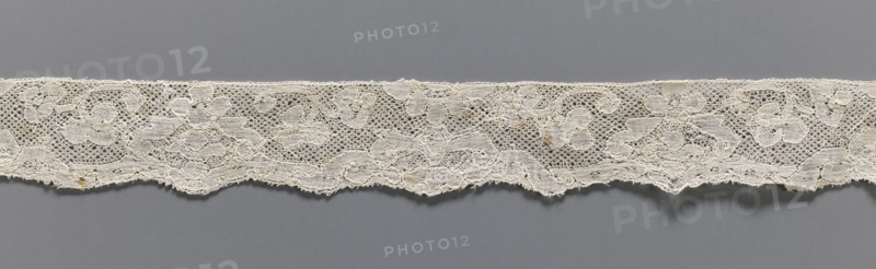 Lace fragment, lace, Malines Mechelen lace, motifs outlined with additional thread, Ißgrond background, lower edge slightly serrated, 2nd half of the 18th century, width 3.5 cm, length 37 cm, A horizontal strip of delicate lace with an irregular scalloped lower edge stretches across a gray background; the lace is a pale off-white or cream color with visible floral and looping mesh patterns and a few small darker spots and slight fraying along the edges.